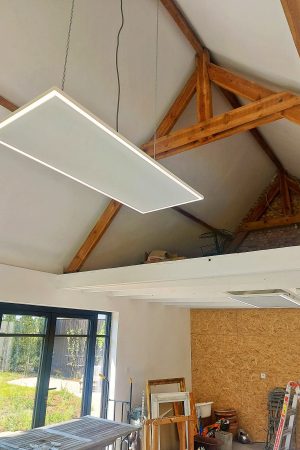 Zwevend infraroodpaneel met LED-verlichting opgehangen aan kettingen onder een hoog dak met houten spanten